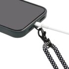 Close-up of a smartphone with a dark case. A charging cable is plugged into the phone, and a black and white lanyard is attached to the case with a clip. The lanyard is partially visible.