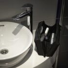 A sleek bathroom sink with a tall, modern faucet is beside a black toiletry bag. The bag is open, displaying a toothbrush, toothpaste, and a can of shaving cream. The setting is minimalistic with a dark countertop and white basin.