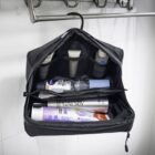 Toiletry bag hanging open, displaying neatly organized items: a deodorant, toothpaste, toothbrush, hair products, and various small containers. The bag has multiple compartments and hangs from a hook in a bathroom setting.