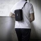 A person wearing a white shirt and dark pants is standing with their back to the camera in a dimly lit room. They have a small black shoulder bag strapped across their back. The wall beside them is textured and appears worn.