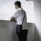 A person with short dark hair stands in a white polo shirt and dark pants, facing away. They lean on a concrete wall, holding a black shoulder bag, in an industrial-looking environment.