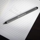 A grey stylus pen labeled PICASSO 2 rests diagonally on an open lined notebook. The page displays handwritten text: HOUSE OF REVOLUTION. The background is a dark, smooth surface.