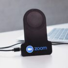 A black wireless charger with the Zoom logo sits on a desk near a laptop. A cable is plugged into the charger, which features a circular charging pad at the top. The surface is white and the background is blurred.
