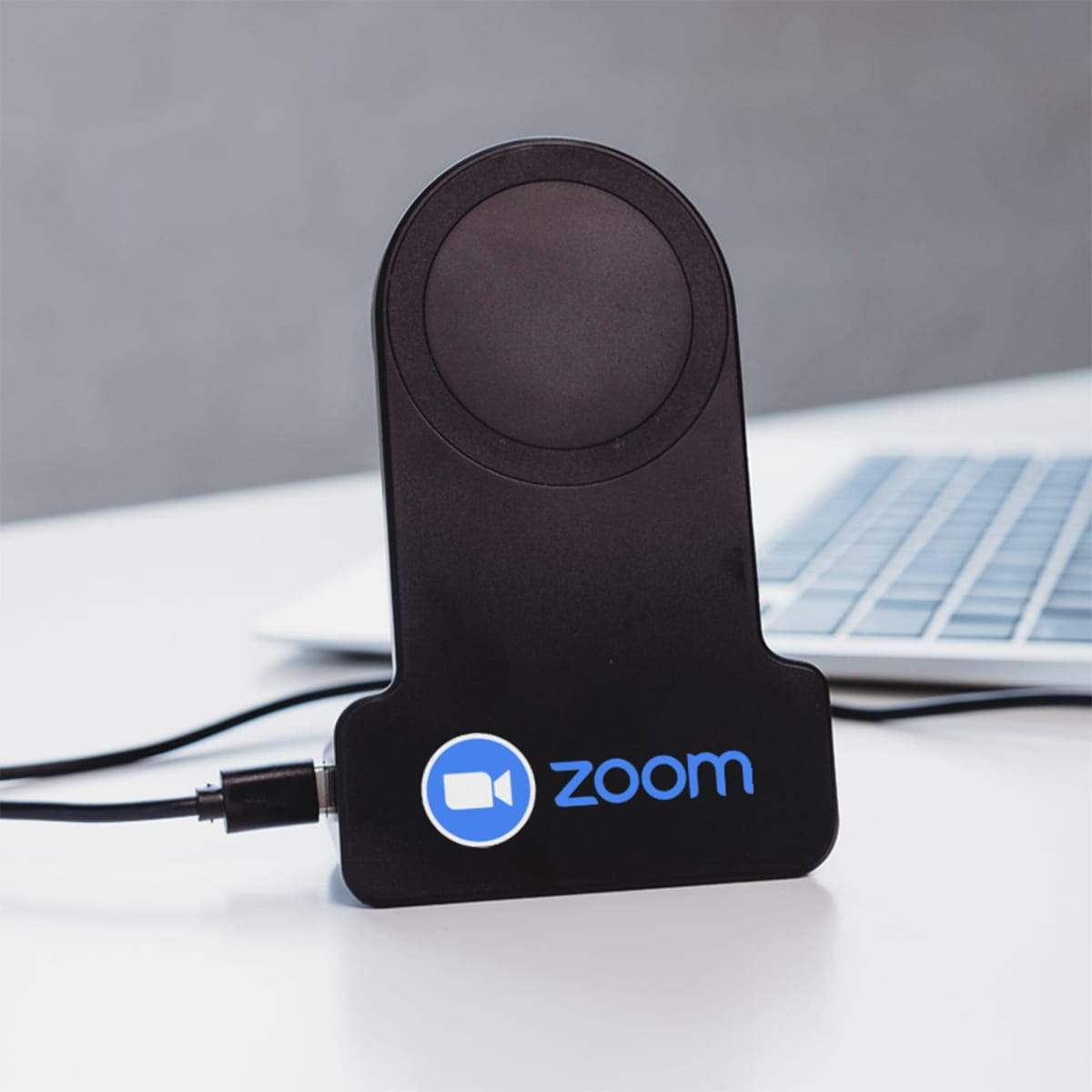 A black wireless charger with the Zoom logo sits on a desk near a laptop. A cable is plugged into the charger, which features a circular charging pad at the top. The surface is white and the background is blurred.