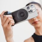 A person holding a smartphone with a circular grip attachment on the back, facing the camera. The focus is on the phones back, showing the lens and attachment, while the person is slightly blurred in the background.