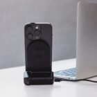 A smartphone with a black case is docked in a charging stand connected to a laptop. The phone is upright, and the laptop is partially visible in the background on a white surface. The setting appears to be a modern workspace.