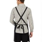 A person with short brown hair, wearing a black and white checkered shirt and black pants, stands with their back turned. They have a black apron tied around the waist, with straps crossing at the back.