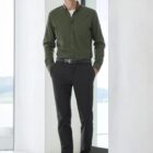 A person stands indoors wearing a green button-up shirt, black pants, and brown shoes. They are next to a large window with a blurred outdoor view.