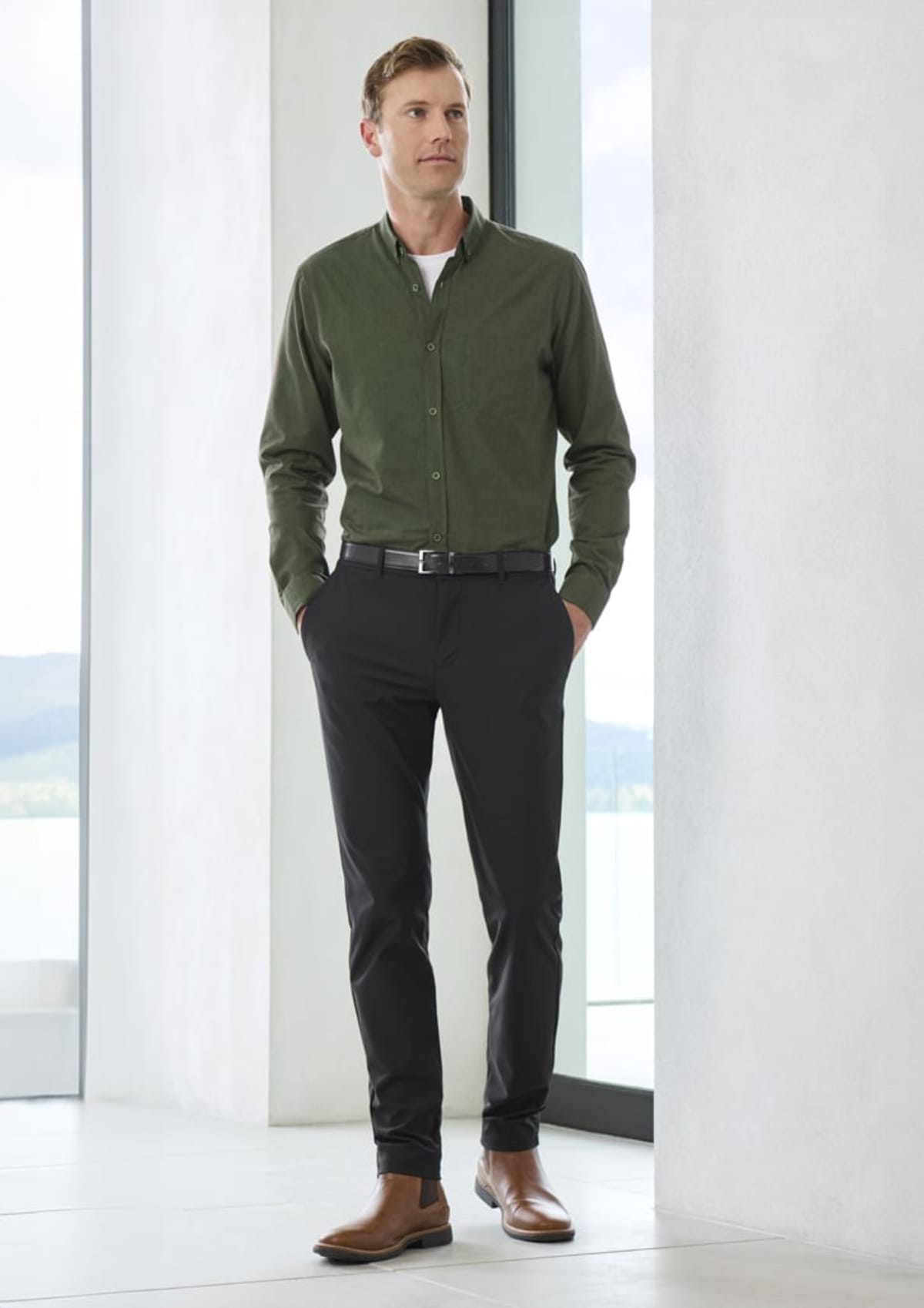 A person stands indoors wearing a green button-up shirt, black pants, and brown shoes. They are next to a large window with a blurred outdoor view.