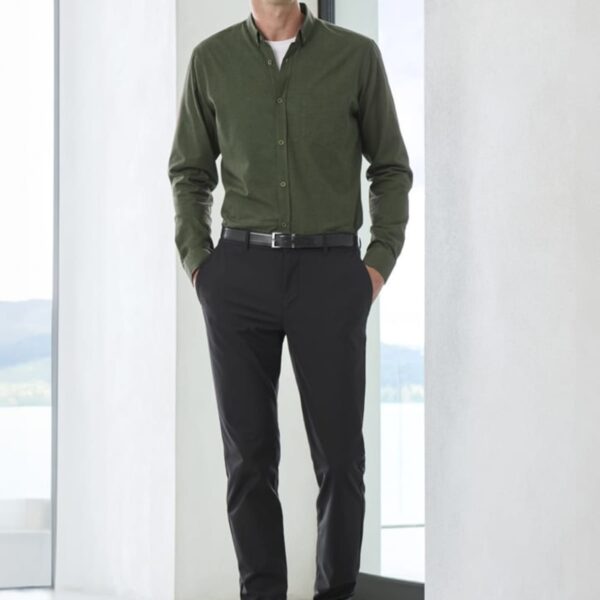 A person stands indoors near large windows, wearing a green button-up shirt, black pants, a black belt, and brown shoes. The setting appears modern, with a light-colored floor and walls.