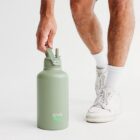 A person wearing white sneakers and socks is picking up a large olive green water bottle with GrabPay written on it. The background is plain white, highlighting the casual, sporty attire and the bottles minimalist design.