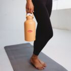 A person in black leggings stands barefoot on a yoga mat, holding a large beige Lipton thermos. The setting appears to be a bright, minimalist room.