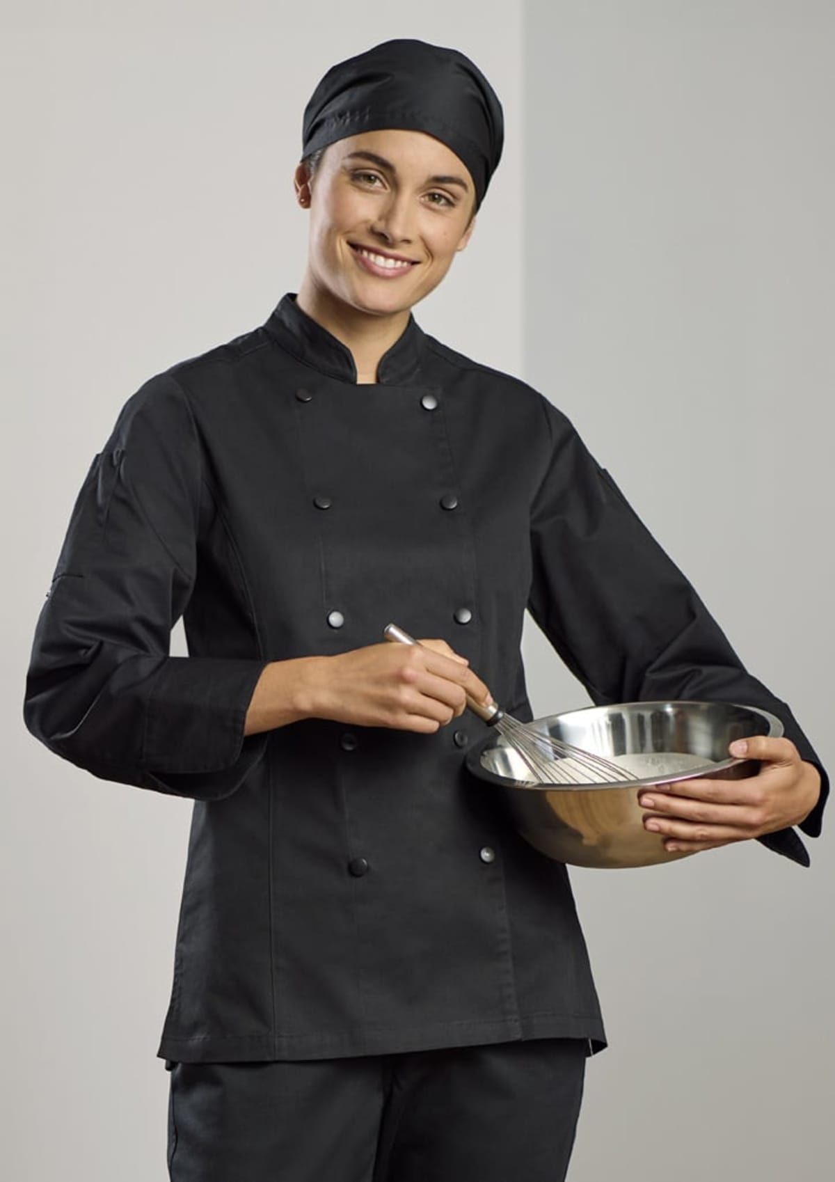 A smiling person in a black chefs uniform and hat is holding a whisk and mixing ingredients in a metal bowl. They are standing in front of a light gray background.