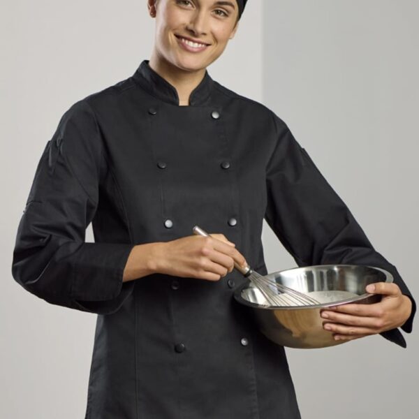 A person in a black chefs uniform and hat is smiling while holding a metal mixing bowl and whisk. They appear to be in a kitchen setting.