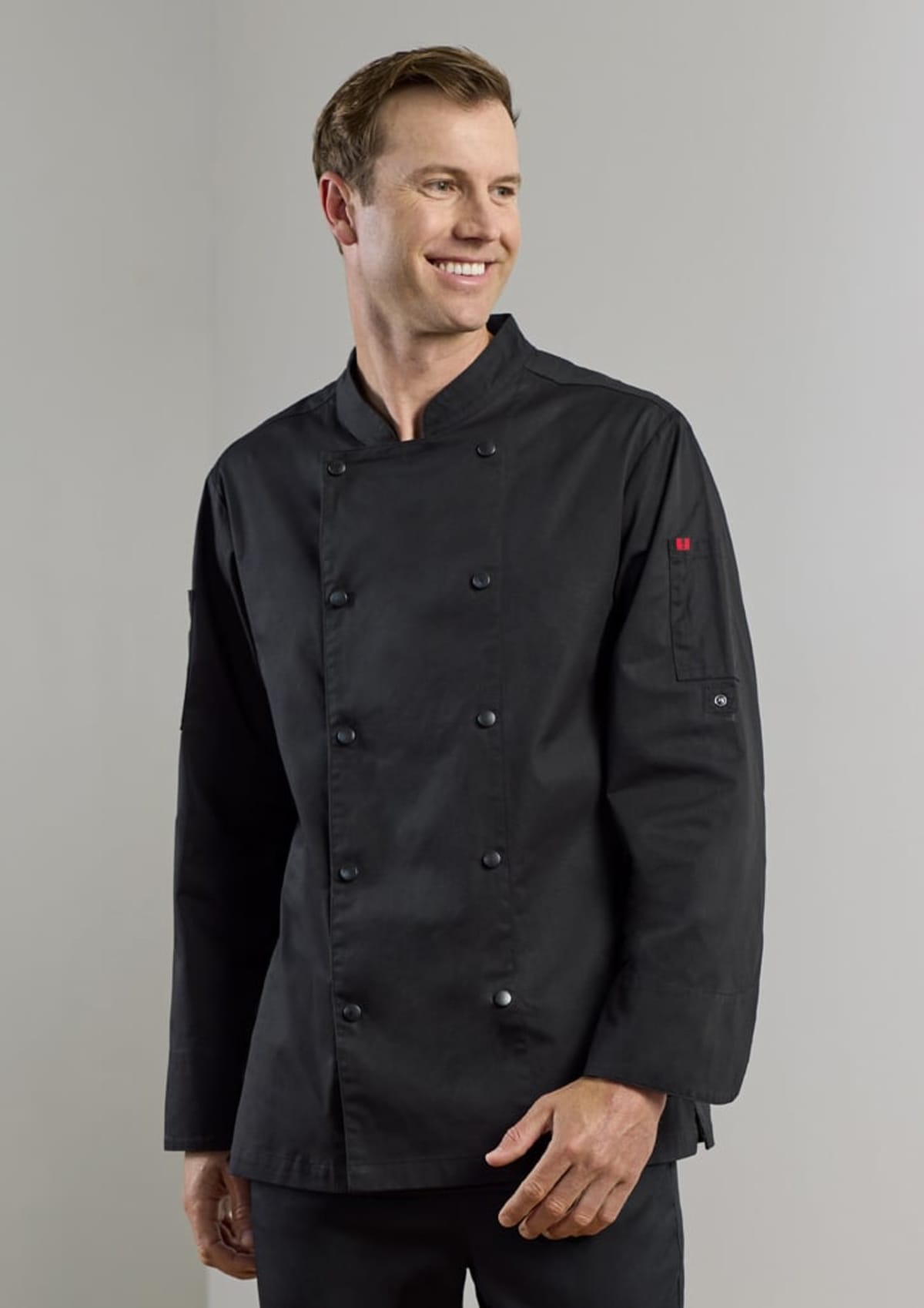 Smiling person wearing a black chefs coat with buttons, standing against a plain background.
