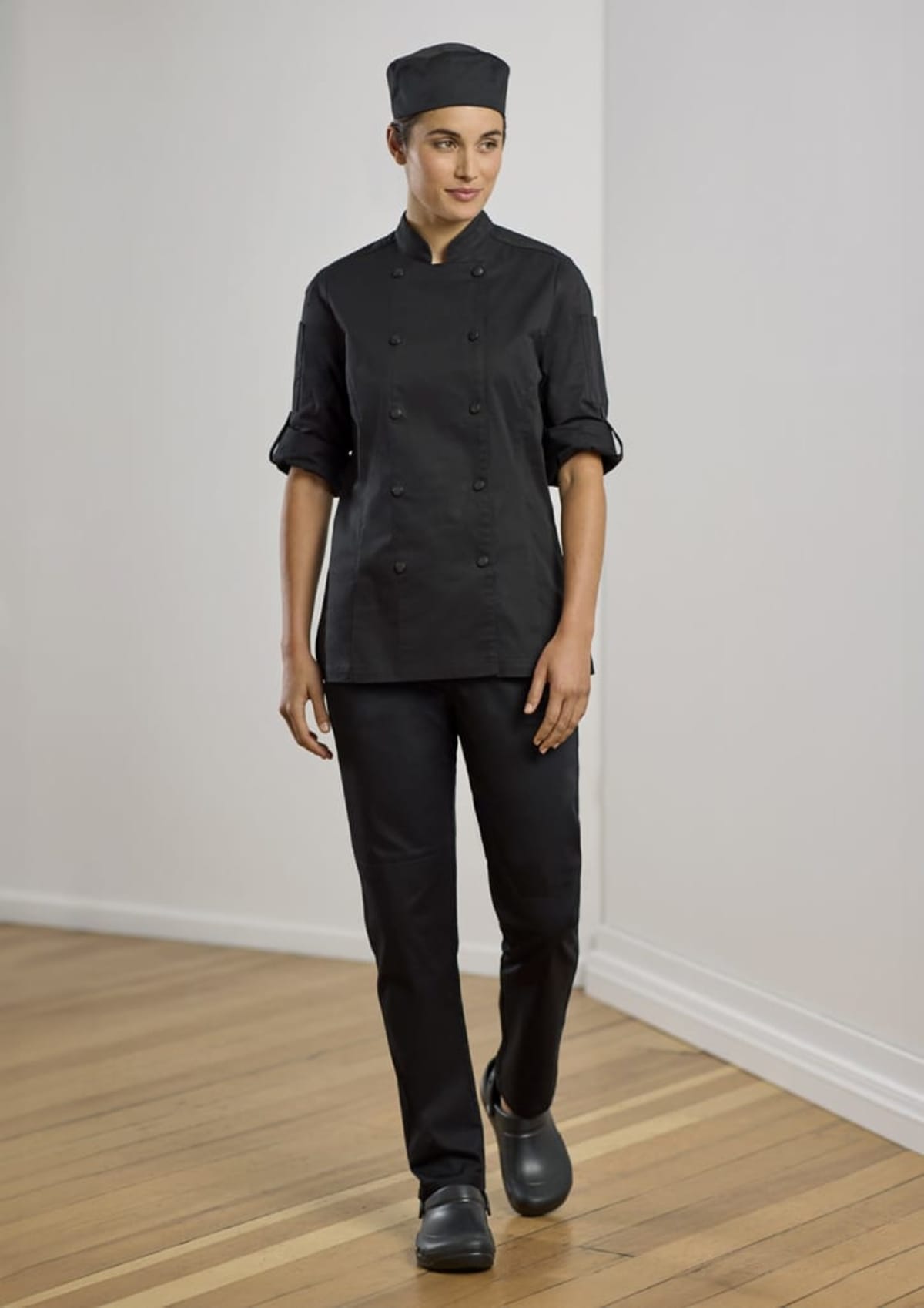 A person wearing a black chef uniform, including a cap, walks on a wooden floor. Their sleeves are rolled up, and they are indoors against a plain wall.