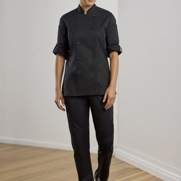 Person walking on a wooden floor, wearing a black chef uniform with a cap, long sleeves rolled up, and black shoes. The background is a plain, light-colored wall.