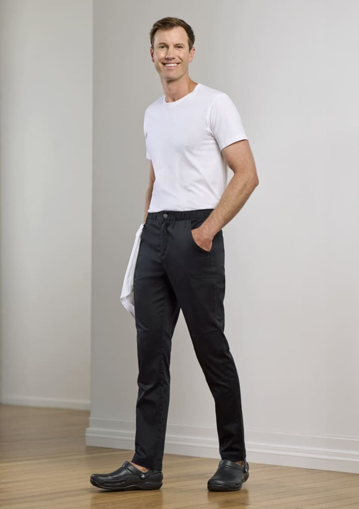 A person stands indoors, smiling and wearing a white T-shirt, dark pants, and black shoes. They hold a white cloth in one hand. The background features a light-colored wall and wooden flooring.