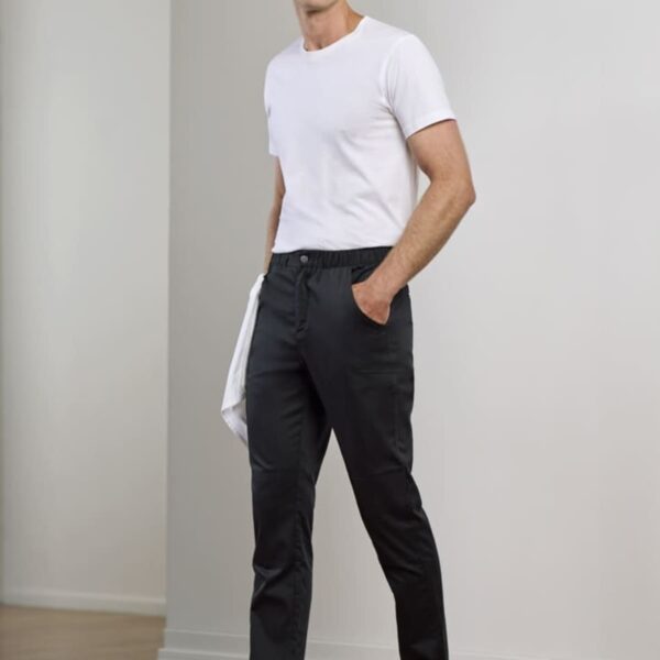 A man in a white T-shirt and black pants stands indoors on a wooden floor, holding a white cloth in one hand. He is smiling and wearing black shoes. The background features a white wall.