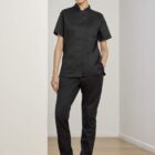 Person standing in a hallway wearing black scrub top and pants with hands in pockets. They are also wearing black clogs and smiling slightly. The walls and floor are light-colored.