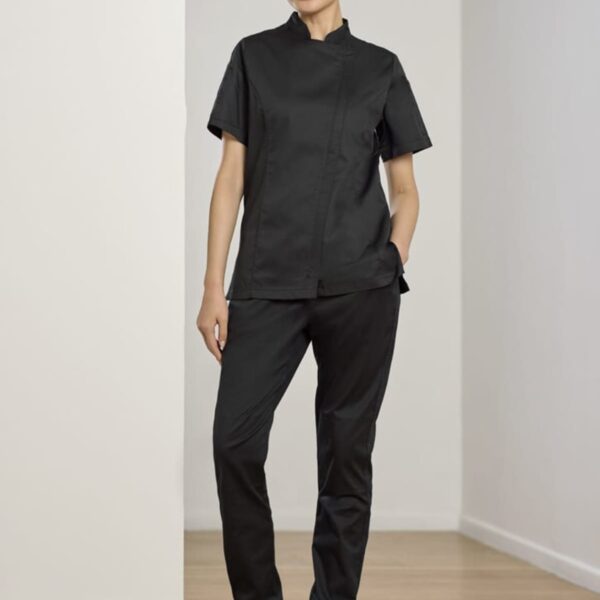 Person standing in a hallway wearing black scrub top and pants with hands in pockets. They are also wearing black clogs and smiling slightly. The walls and floor are light-colored.