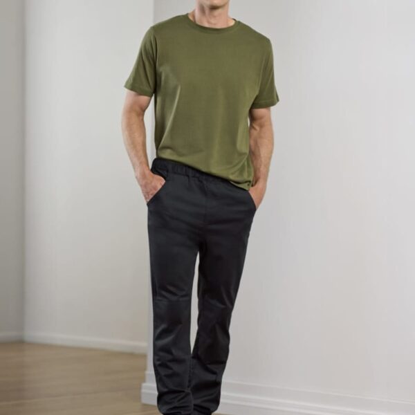A person stands indoors on a wooden floor. They are wearing a green T-shirt, black jogger pants, and black sneakers. Their hands are in their pockets, and they are looking at the camera. The background is a plain, light-colored wall.