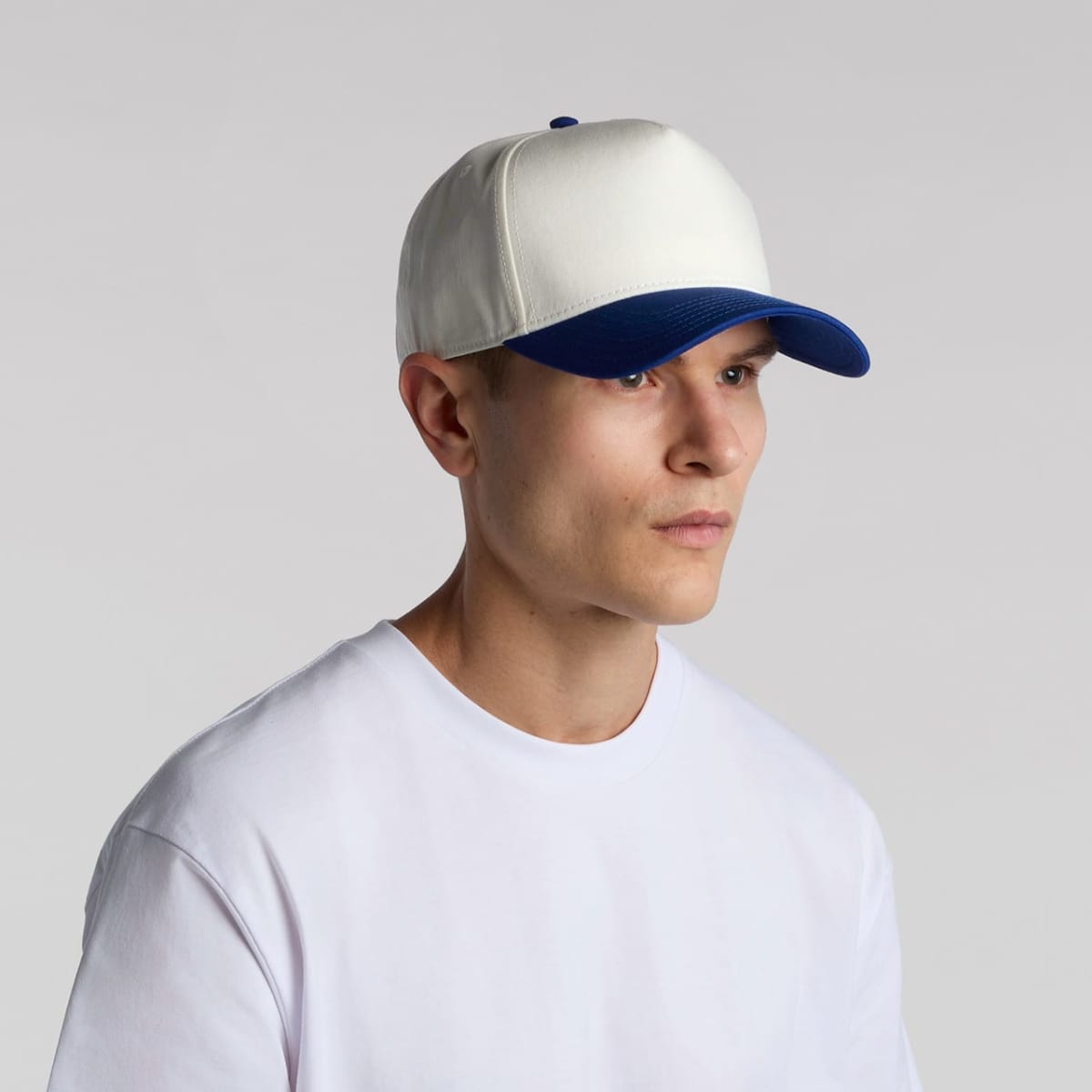 A person wearing a white baseball cap with a blue brim and a white t-shirt, looking to the side against a plain gray background.