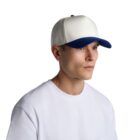 A person wearing a white t-shirt and a white baseball cap with a blue brim, looking to the left. The background is plain white.