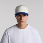 A person wearing a plain white t-shirt and a white cap with a blue brim is standing against a neutral gray background.