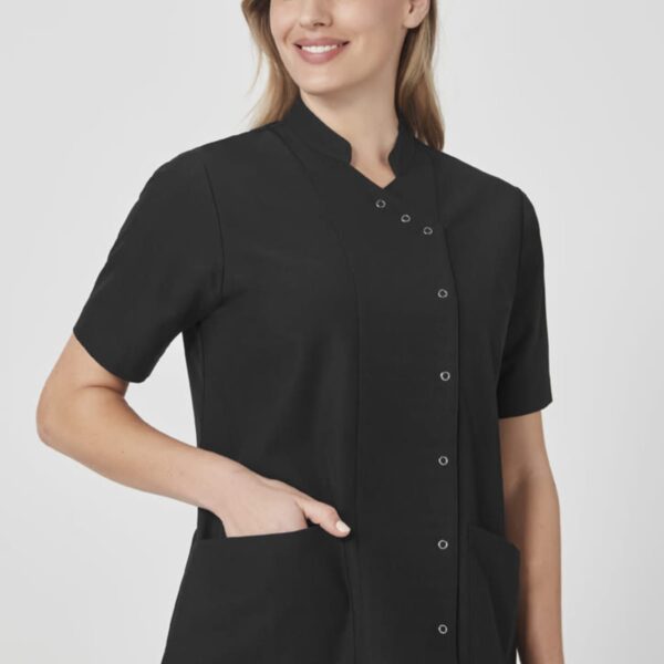 A person wearing a short-sleeved, black button-up uniform top with front pockets, smiles while looking to the side. They have long, light brown hair and are posing against a plain background.