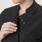 A person wearing a black garment with silver snap buttons is partially visible from the shoulders to the mouth. They are holding the edge of the garment with one hand, which has neatly manicured nails. The background is a neutral light color.