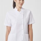 A person wearing a white short-sleeved, button-up uniform with two pockets stands against a plain background. The person is smiling and looking slightly to the side.