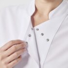 A person wearing a white chefs coat with metal snap buttons is adjusting the collar. The coat features a high neck and the persons hand is visible, interacting with the garment, showcasing the details of the buttons.