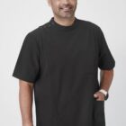 A person with dark hair and a beard is smiling and wearing a short-sleeved black uniform with a pocket. They have a watch on their left wrist and are standing against a plain white background.