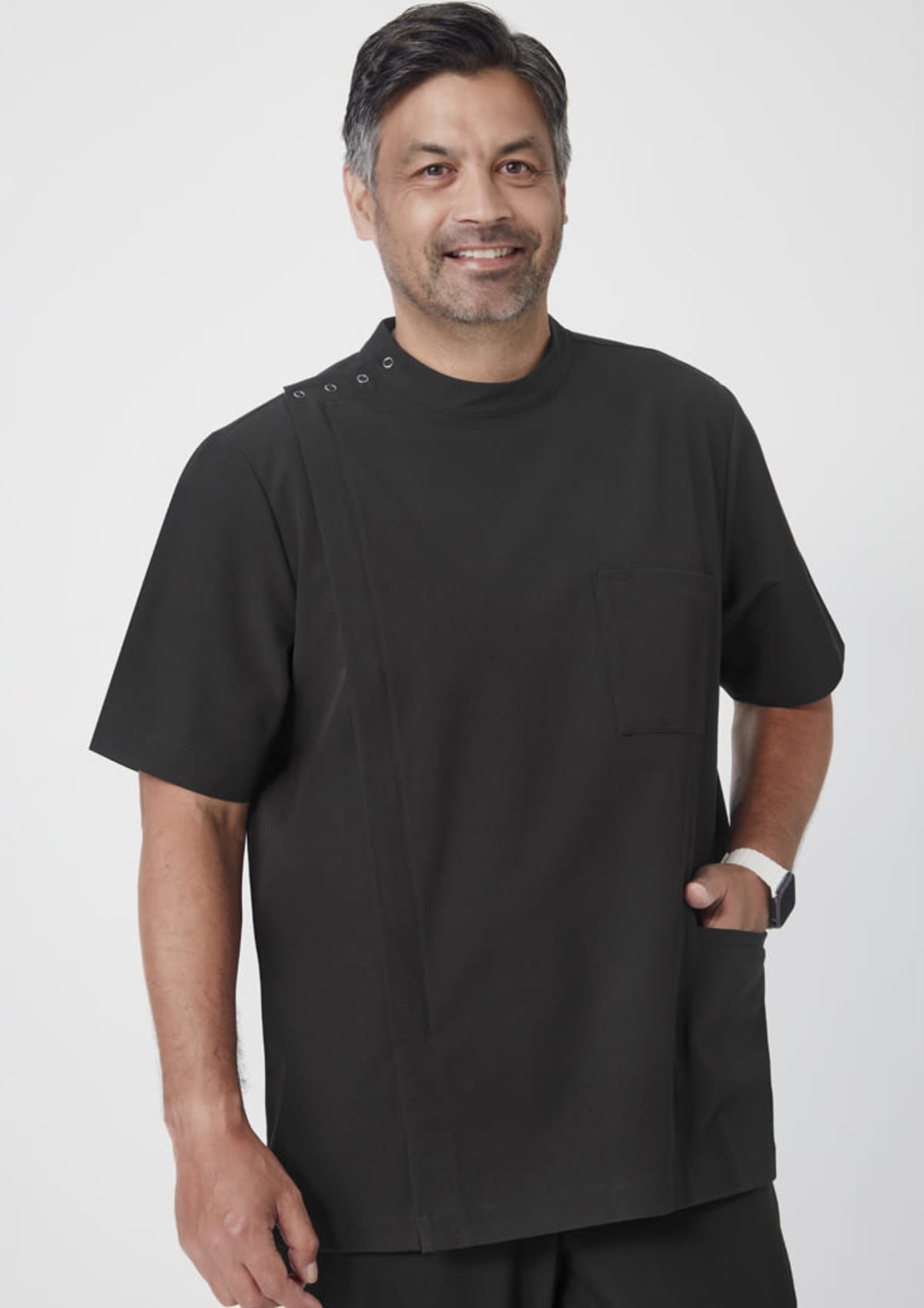 A person with dark hair and a beard is smiling and wearing a short-sleeved black uniform with a pocket. They have a watch on their left wrist and are standing against a plain white background.