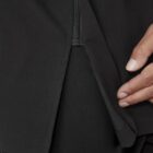 A close-up of a black garment with a discreet zippered pocket. A persons hand is partially visible, holding the edge of the fabric near the pocket opening. The material appears smooth and slightly stretchy.