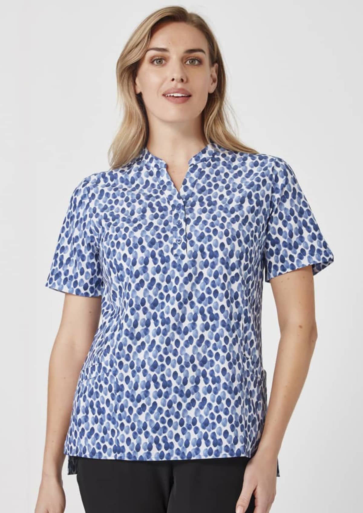 A person wearing a blue and white dotted blouse with short sleeves and a collarless V-neck, standing against a plain white background.