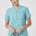 A person in a blue and white patterned scrub top stands against a plain background, smiling gently. They hold a stethoscope in their hands and are wearing black pants.