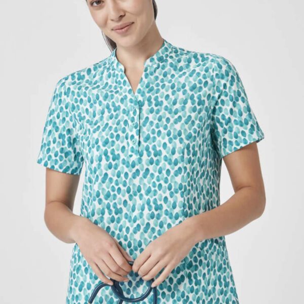 A person in a blue and white patterned scrub top stands against a plain background, smiling gently. They hold a stethoscope in their hands and are wearing black pants.