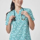A person with long hair smiles while wearing a blue and white patterned medical scrub top and holding a stethoscope around their neck. The background is plain and light-colored.