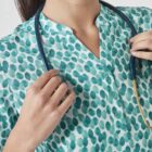 A person wearing a teal and white polka-dot shirt holds a stethoscope around their neck. The image focuses on the upper body, with the individuals hands visible holding the stethoscope.
