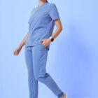 A person wearing light blue scrubs and gray sneakers stands against a light blue background. They have dark hair tied back and wear a smartwatch. The outfit consists of a short-sleeved top and pants with pockets.
