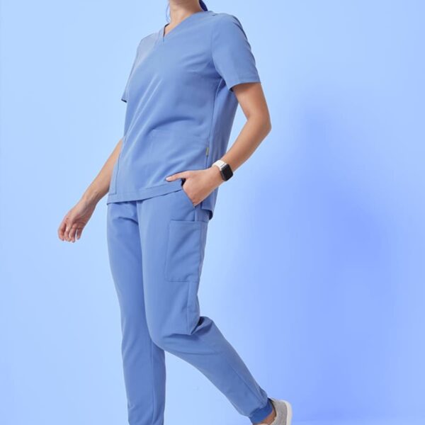 A person wearing light blue scrubs and gray sneakers stands against a light blue background. They have dark hair tied back and wear a smartwatch. The outfit consists of a short-sleeved top and pants with pockets.