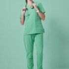 A person in green medical scrubs stands on a matching green background. They have a stethoscope around their neck and are looking upwards to the right, with hands resting in their pockets. Gray sneakers complete the outfit.