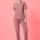 A person stands against a solid pink background wearing a pink scrub top and pants with gray sneakers. They have dark hair pulled back and wear a watch. The outfit appears comfortable and practical, suitable for a healthcare setting.