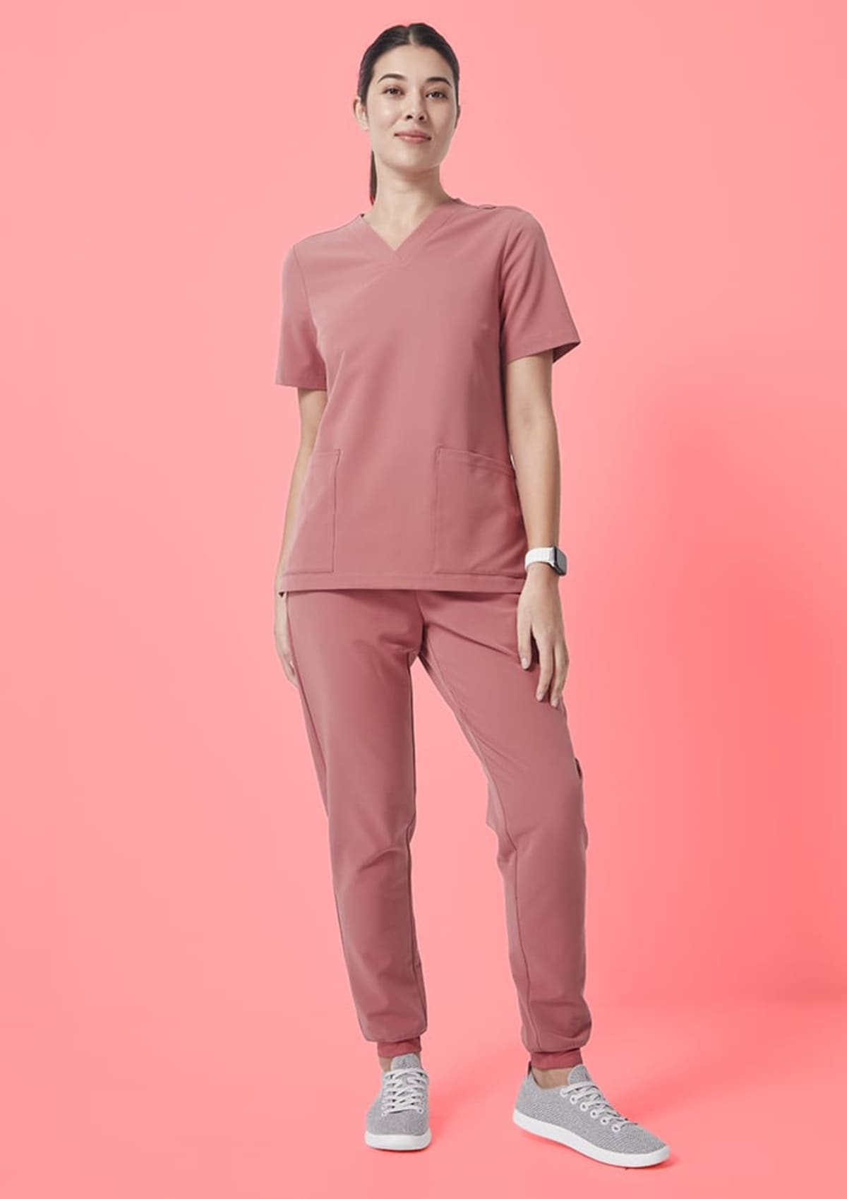 A person stands against a solid pink background wearing a pink scrub top and pants with gray sneakers. They have dark hair pulled back and wear a watch. The outfit appears comfortable and practical, suitable for a healthcare setting.