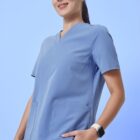 A person wearing blue scrubs stands against a blue background. They are looking slightly upwards, with one hand in their pocket. They are also wearing a smartwatch on their wrist.
