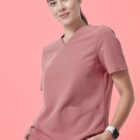 A person with dark hair pulled back wears a pink medical scrub top and pants, standing against a pink background. They have one hand in a pocket and a smartwatch on their wrist, smiling gently at the camera.