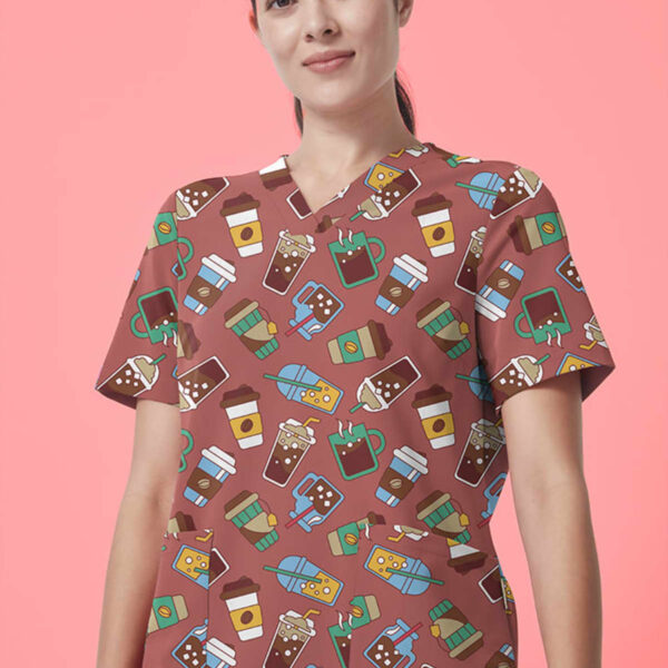 A person wearing scrubs with a coffee-themed pattern stands against a pink background. The scrubs are adorned with illustrations of coffee cups, to-go cups, and coffee beans. The person has long, dark hair tied back and is wearing a wristband.
