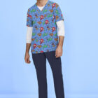 A man is standing against a light blue background, wearing colorful scrubs featuring cartoon characters and dinosaurs. He has a long-sleeve white shirt underneath and is also wearing navy blue pants and gray sneakers.